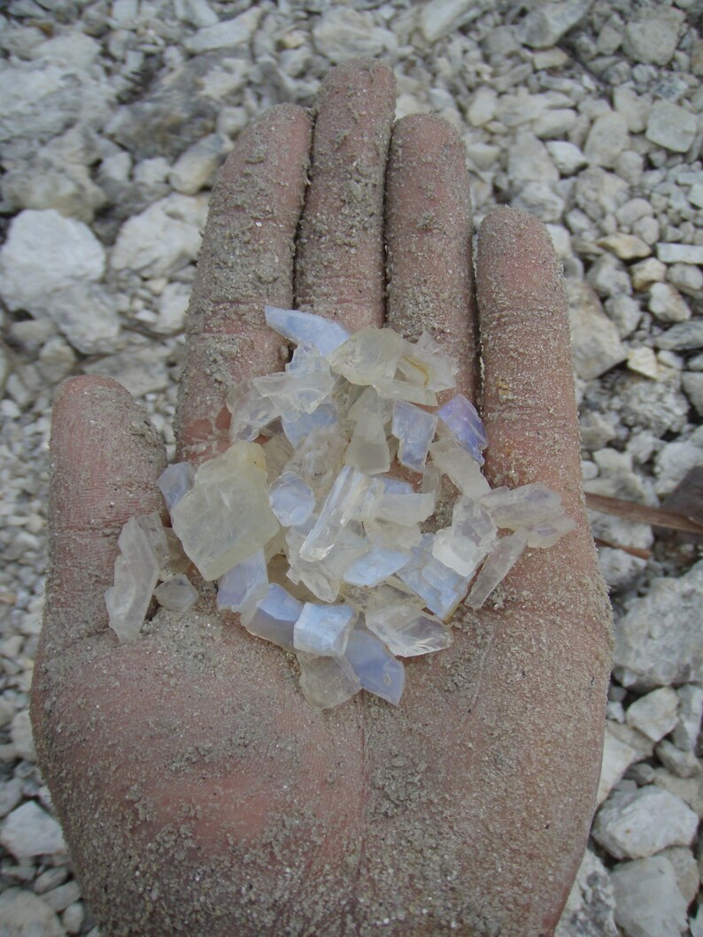 May include: A collection of small, translucent, and semi-opaque raw mineral fragments in shades of pale blue, white, and clear. The stones have irregular, jagged edges and a smooth, glassy texture, appearing as unpolished, natural crystal pieces.