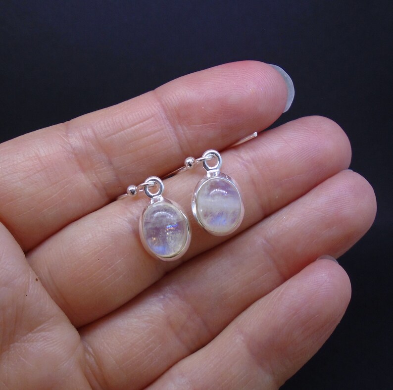 May include: A pair of oval-shaped moonstone earrings set in polished silver bezels. Each earring features a smooth, translucent stone with a subtle iridescent blue sheen, suspended from a simple silver hook wire for a clean, classic look.
