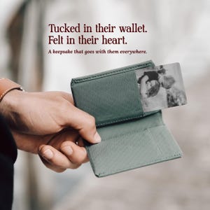 May include: A sage green wallet featuring a diagonal textured pattern and a rectangular metal insert displaying a black and white photograph of two children. The text reads: Tucked in their wallet. Felt in their heart. A keepsake that goes with them everywhere.
