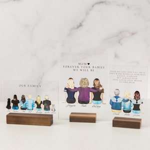 May include: Two acrylic plaques with illustrations of families. The top plaque reads "Mom Forever Your Babies We Will Be" and features three women with the names "Shagna", "Thom", and "Kelsey" written below them. The bottom plaque reads "Our Family" and features seven people with a dog and a cat. The names "Erin", "Madison", "Dad", "Brody", "Garret", and "Gwen" are written below the figures.