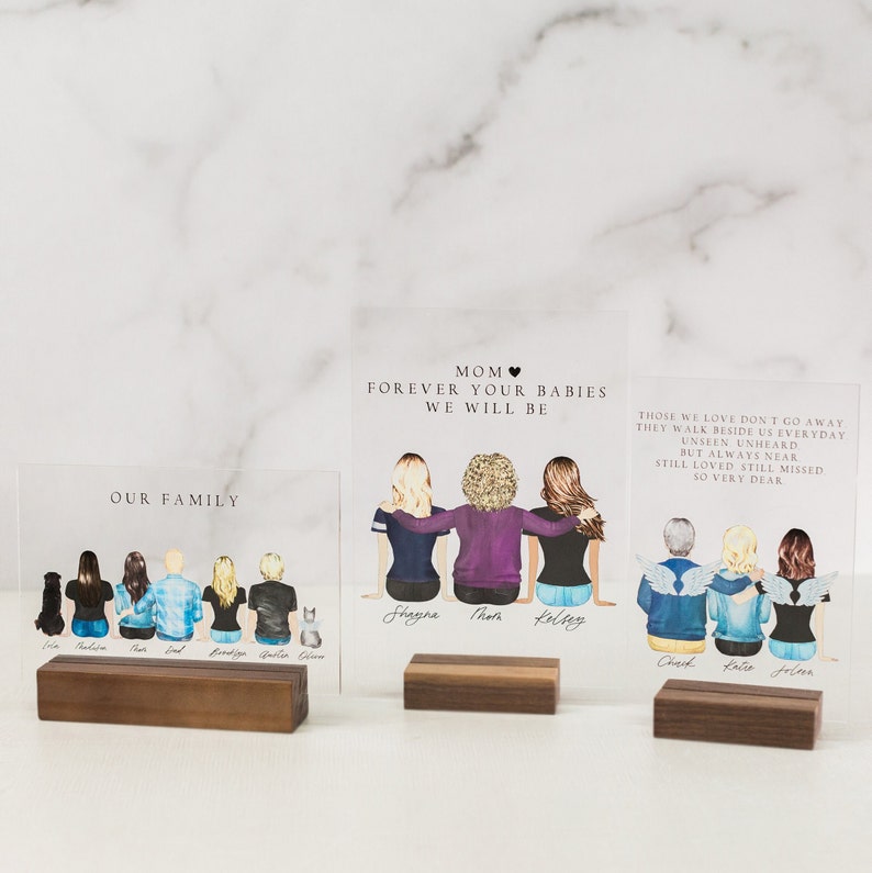 May include: Two acrylic plaques with illustrations of families. The top plaque reads "Mom Forever Your Babies We Will Be" and features three women with the names "Shagna", "Thom", and "Kelsey" written below them. The bottom plaque reads "Our Family" and features seven people with a dog and a cat. The names "Erin", "Madison", "Dad", "Brody", "Garret", and "Gwen" are written below the figures.