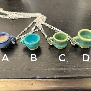 May include: Four miniature ceramic teacup pendants on silver chains. The teacups are in shades of blue and green, labeled A, B, C, and D. Each cup has a handle and a unique glaze. The necklaces are displayed on a dark surface.