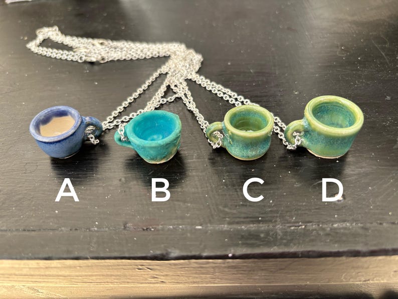 May include: Four miniature ceramic teacup pendants on silver chains. The teacups are in shades of blue and green, labeled A, B, C, and D. Each cup has a handle and a unique glaze. The necklaces are displayed on a dark surface.