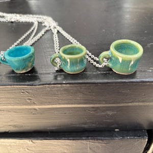 May include: Miniature ceramic teacup necklaces in shades of blue and green. Each tiny cup is attached to a silver chain. The cups have a rustic, handmade appearance, with visible texture and variations in color. The necklaces are displayed on a dark surface.