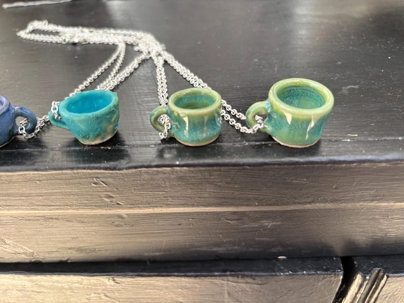 May include: Miniature ceramic teacup necklaces in shades of blue and green. Each tiny cup is attached to a silver chain. The cups have a rustic, handmade appearance, with visible texture and variations in color. The necklaces are displayed on a dark surface.