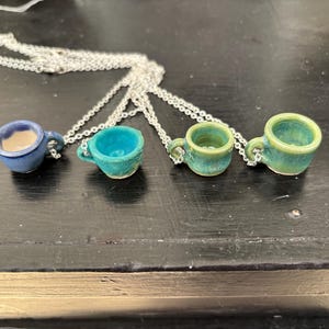 May include: Four miniature ceramic teacup pendants on silver chains. The teacups are in shades of blue and green, with visible textures and small handles. The necklaces are displayed on a dark surface.