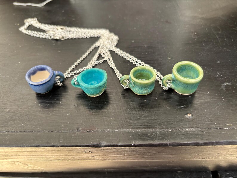 May include: Four miniature ceramic teacup pendants on silver chains. The teacups are in shades of blue and green, with visible textures and small handles. The necklaces are displayed on a dark surface.
