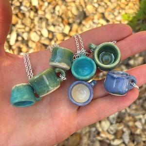 May include: A collection of miniature ceramic mug pendants in various shades of blue and green. Each tiny mug is attached to a silver chain. The pendants are held in a hand, showcasing the different designs and colors of the miniature mugs.