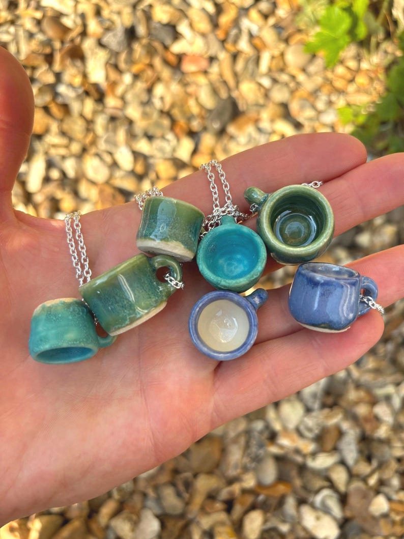May include: A collection of miniature ceramic mug pendants in various shades of blue and green. Each tiny mug is attached to a silver chain. The pendants are held in a hand, showcasing the different designs and colors of the miniature mugs.