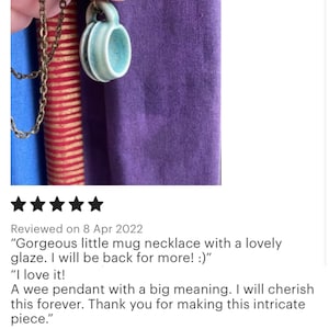 May include: A small, light blue ceramic mug pendant with a handle, hanging on a gold-colored chain. The mug has a textured surface and is set against a purple background. The pendant is a miniature version of a coffee mug.