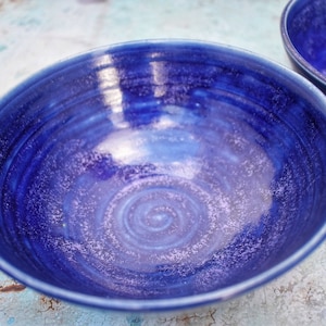Może przedstawiać: Dwie ręcznie robione ceramiczne miski z głębokim niebieskim szkliwem. Miski mają spiralny wzór na środku i błyszczące wykończenie. Miski są w żywym odcieniu niebieskiego, idealne do serwowania jedzenia.