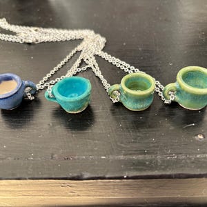 May include: Four miniature ceramic teacup pendants in shades of blue and green hang from silver chains. Each cup has a small handle and a unique glaze. The necklaces are displayed on a dark surface, showcasing the handcrafted details.