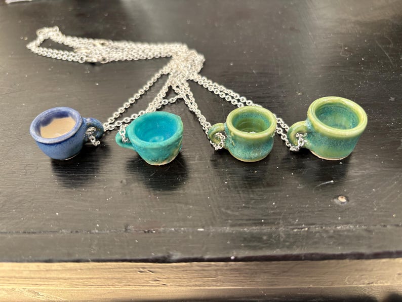 May include: Four miniature ceramic teacup pendants in shades of blue and green hang from silver chains. Each cup has a small handle and a unique glaze. The necklaces are displayed on a dark surface, showcasing the handcrafted details.