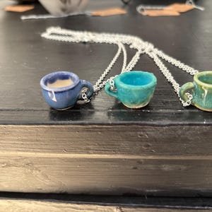 May include: Three ceramic coffee cup pendants on silver chains. The cups are in shades of blue, teal, and green. The blue cup has a creamy interior, while the others are empty. Each cup has a small handle.