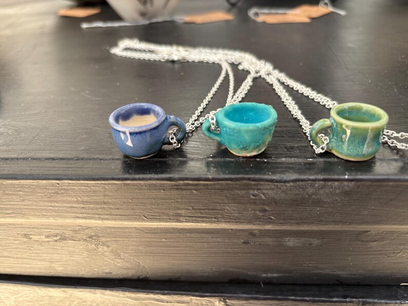 May include: Three ceramic coffee cup pendants on silver chains. The cups are in shades of blue, teal, and green. The blue cup has a creamy interior, while the others are empty. Each cup has a small handle.