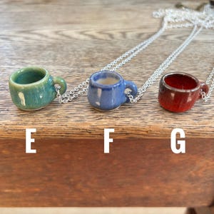 May include: Three miniature ceramic mug pendants in teal, blue, and red, each suspended from a silver chain. The mugs have small handles and are displayed on a wooden surface. The letters "E", "F", and "G" are printed below each mug.