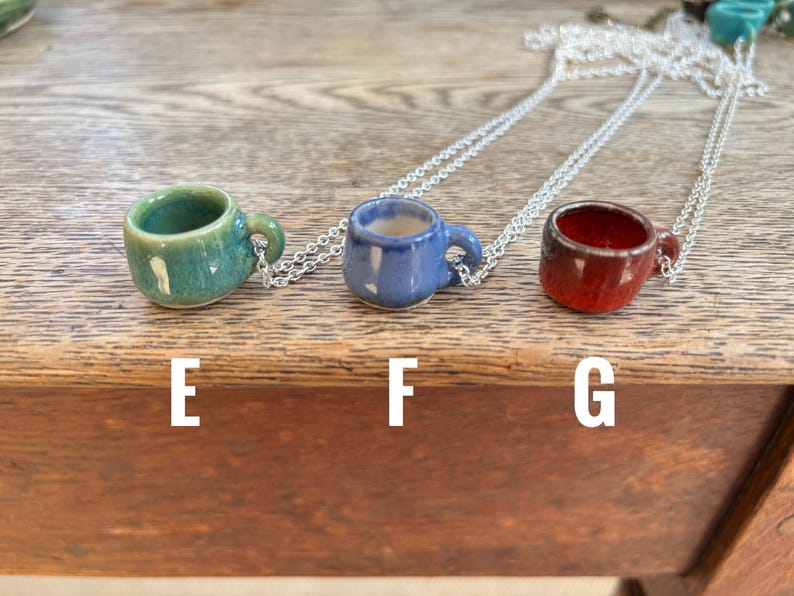 May include: Three miniature ceramic mug pendants in teal, blue, and red, each suspended from a silver chain. The mugs have small handles and are displayed on a wooden surface. The letters "E", "F", and "G" are printed below each mug.