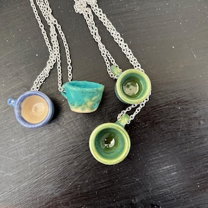 May include: Four ceramic cup necklaces with silver chains. The cups are in shades of blue, green, and turquoise, with a small handle on each. The cups have a glossy finish and are arranged on a dark surface.
