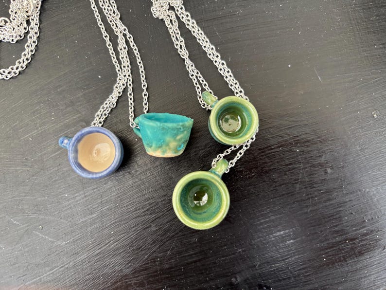 May include: Four ceramic cup necklaces with silver chains. The cups are in shades of blue, green, and turquoise, with a small handle on each. The cups have a glossy finish and are arranged on a dark surface.