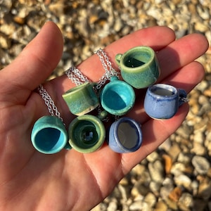 May include: A collection of miniature ceramic mug pendants in various shades of blue and green, each suspended from a delicate silver chain. The tiny mugs have a glossy finish and are held in a hand against a blurred background.