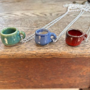May include: Three miniature ceramic coffee cup pendants in teal, blue, and red, each suspended from a silver chain. The cups have small handles and are displayed on a wooden surface. The necklaces are a unique accessory.