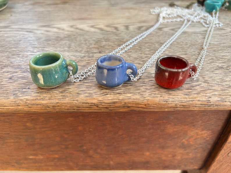 May include: Three miniature ceramic coffee cup pendants in teal, blue, and red, each suspended from a silver chain. The cups have small handles and are displayed on a wooden surface. The necklaces are a unique accessory.