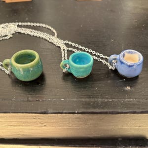 May include: Three miniature ceramic coffee cup pendants on silver chains. The cups are in shades of green, teal, and blue. Each cup has a small handle and is attached to a delicate chain, creating a unique jewelry piece.