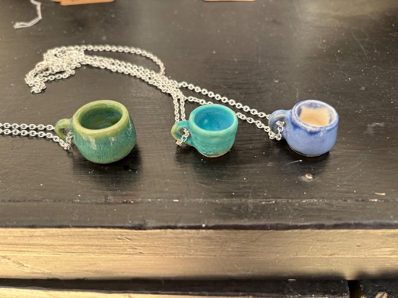 May include: Three miniature ceramic coffee cup pendants on silver chains. The cups are in shades of green, teal, and blue. Each cup has a small handle and is attached to a delicate chain, creating a unique jewelry piece.