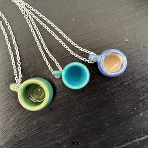 May include: Three ceramic cup pendants on silver chains. The cups are in shades of green, teal, and blue, with a brown interior in one. The pendants are small, with a loop for the chain. The background is a dark, textured surface.
