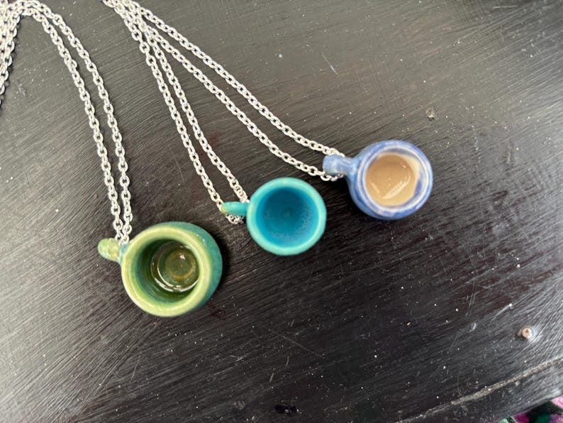 May include: Three ceramic cup pendants on silver chains. The cups are in shades of green, teal, and blue, with a brown interior in one. The pendants are small, with a loop for the chain. The background is a dark, textured surface.