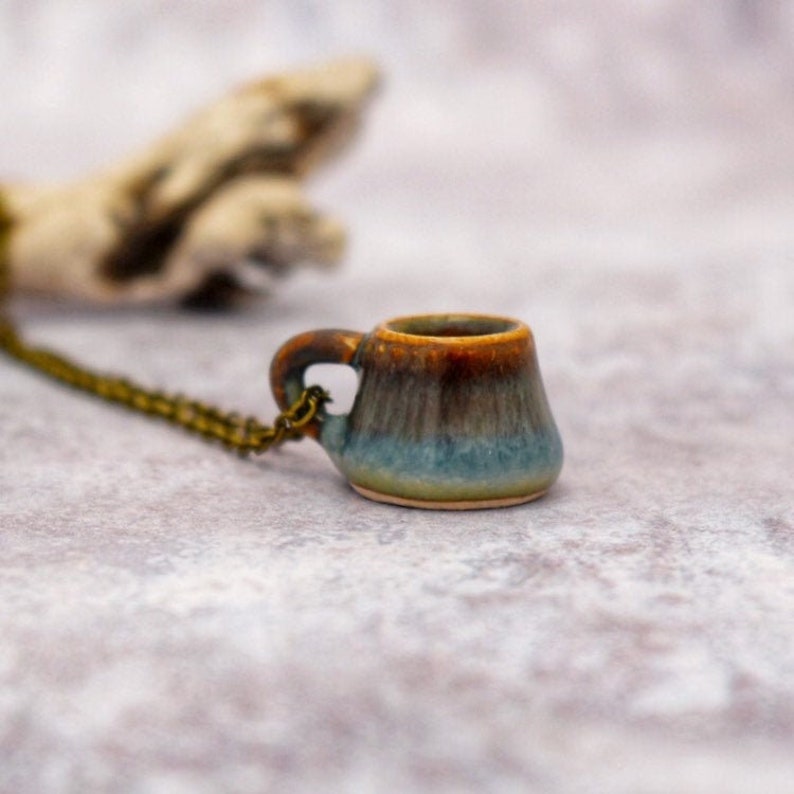 May include: A miniature ceramic mug pendant with a bronze-colored chain. The mug features a gradient of colors, including brown, blue, and tan. The small handle is attached to the side of the mug. The pendant is a unique piece of jewelry.