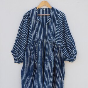 May include: A midi-length dress in deep indigo blue with a white vertical striped pattern. The garment features three-quarter length sleeves, a gathered waistline, and a split V-neckline with a small collar, hanging on a wooden hanger.