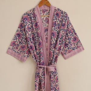 May include: A floral print robe in shades of purple and blue on a cream background. The robe features a striped border and matching belt. The design includes large stylized flowers and leaves. The robe is hanging on a wooden hanger.