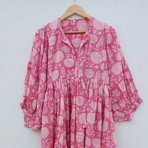 以下が含まれることがあります： 白い花柄のピンクの綿のドレス。ドレスはVネック、七分袖、ギャザーの入ったスカートです。