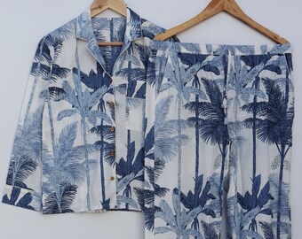 Puede incluir: Un conjunto de pijama blanco con estampado de palmeras azules. La parte superior tiene cuello y mangas largas. Los pantalones tienen cintura con cordón y pierna ancha.