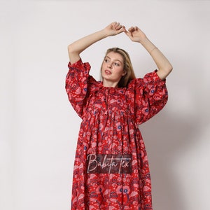 Puede incluir: Un vestido maxi rojo con estampado floral y cintura fruncida. El vestido tiene un escote redondo y mangas 3/4. La modelo lleva sandalias de tac&oacute;n transparentes.