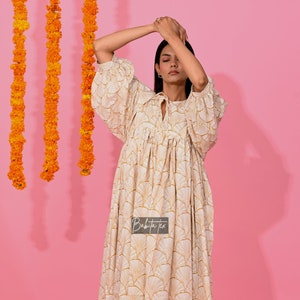Könnte beinhalten: Ein weißes Kleid mit einem goldenen geometrischen Muster. Das Kleid hat eine geraffte Taille und einen leicht bauschigen Ärmel. Das Kleid wird auf einem Model vor einem rosa Hintergrund gezeigt.