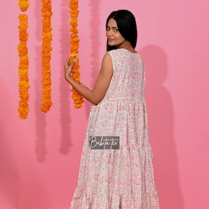 Peut inclure: Une robe maxi blanche sans manches avec un imprimé floral rose. La robe a une jupe à volants et un col rond. Le mannequin porte la robe avec des sandales blanches.