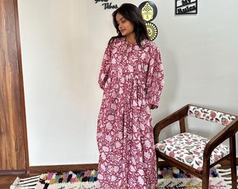 Puede incluir: Una mujer lleva un vestido largo de estampado floral rosa y blanco con falda fruncida. El vestido tiene un escote redondo y mangas largas. La mujer está de pie en una habitación con una pared blanca y una silla de madera con un cojín estampado floral. La pared tiene un letrero de madera que dice "This is our happy place" y otro letrero que dice "My room my rules".