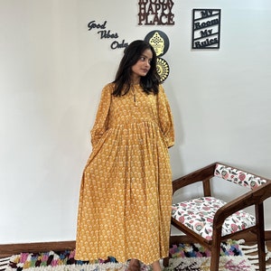 Könnte beinhalten: Eine Frau trägt ein langes, gelbes Blumenkleid mit geraffter Taille. Das Kleid hat einen V-Ausschnitt und lange Ärmel. Die Frau steht vor einer weißen Wand mit einem Holzschild, auf dem "This is our happy place" steht, und einem weiteren Schild, auf dem "My room my rules" steht.