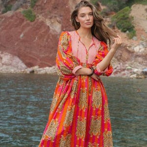 Puede incluir: Un vestido vibrante de manga larga con escote en V, con un llamativo estampado de rayas de tigre en tonos naranja, rosa y marr&oacute;n. El vestido tiene un corpi&ntilde;o a rayas y el texto "BABITAS TEX" en la parte inferior. Se usa al aire libre cerca del agua.