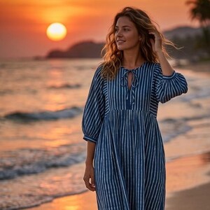 May include: A midi-length dress featuring a dark blue and white vertical striped pattern, three-quarter length sleeves with gathered cuffs, and a round neckline with a central tie closure. The garment has a relaxed fit with a gathered waistline.