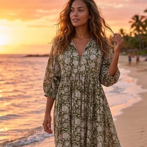 May include: An olive green midi dress with a white floral design. The dress features a V-neck and three-quarter sleeves. The model is on a sandy beach at sunset. The dress is a relaxed fit.