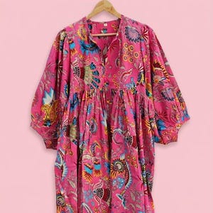 May include: A vibrant pink midi dress featuring a colorful floral and paisley print in shades of blue, yellow, and orange. The dress has a loose, gathered silhouette, a button-up neckline, and voluminous three-quarter length sleeves with elastic cuffs.