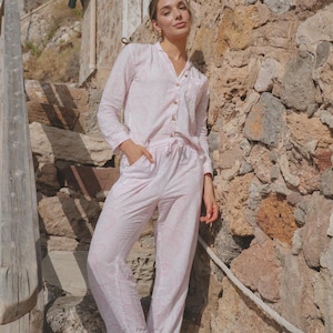 Puede incluir: Un conjunto de pijama rosa claro con un sutil estampado floral. El conjunto incluye una camisa con botones y pantalones a juego. La modelo está de pie sobre escalones de piedra junto a una pared de piedra. El conjunto de pijama es de tela suave y ligera.