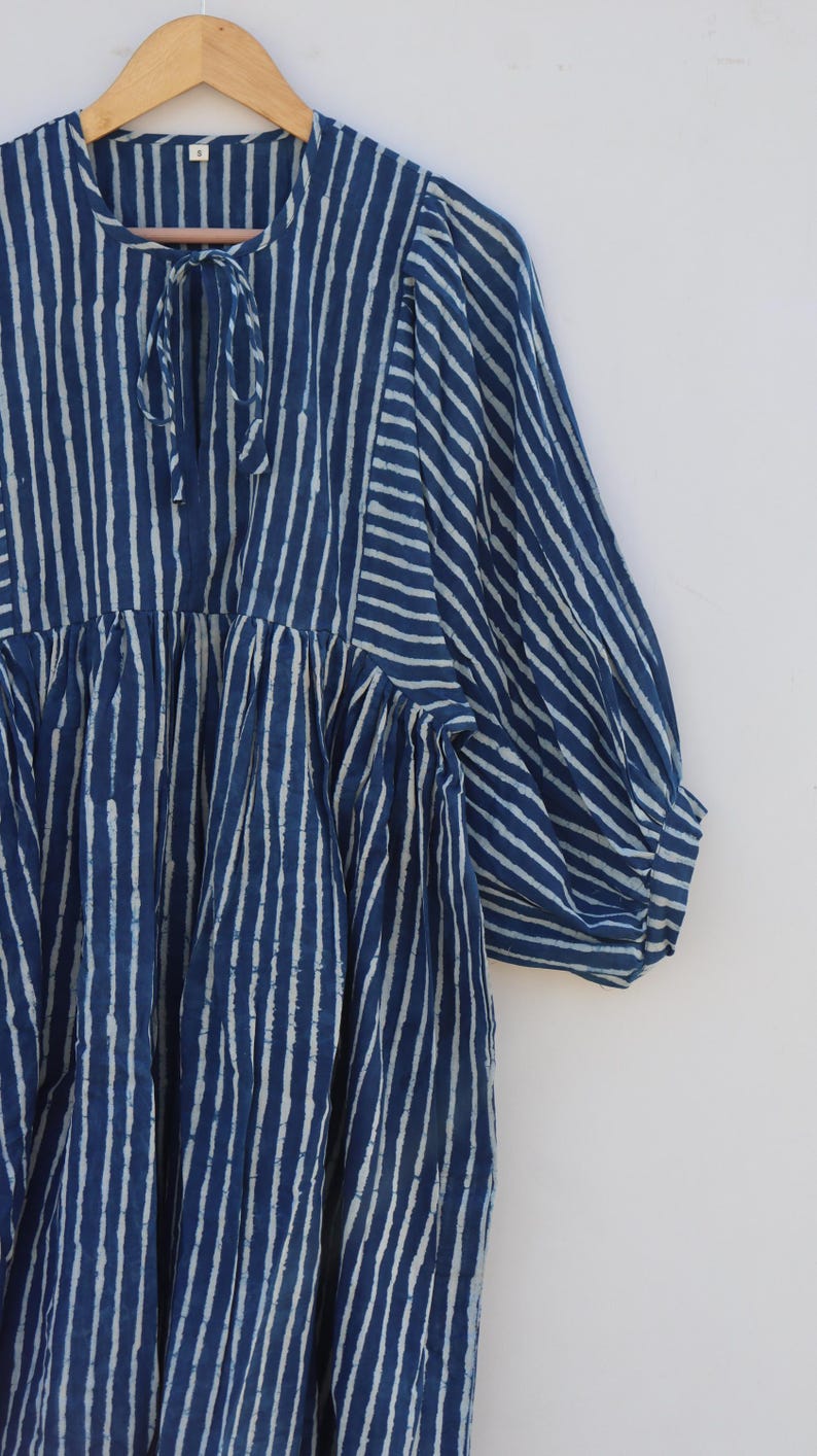 May include: A navy blue and white striped dress featuring a round neckline with a tie closure, gathered fabric at the bodice, and three-quarter length puff sleeves. The garment displays a vertical stripe pattern throughout with a contrasting horizontal stripe detail on the sleeves.