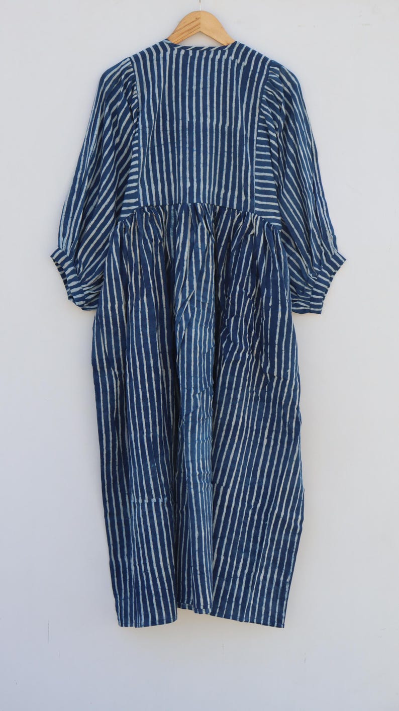 May include: A midi-length dress in deep indigo blue with a white vertical stripe pattern. The garment features three-quarter length sleeves with gathered cuffs, a round neckline, and a gathered waistline that creates a full, flowing skirt silhouette.