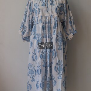 May include: A white cotton dress with a blue floral print and a tie at the neck. The dress has a gathered waist and 3/4 sleeves. The dress is on a mannequin.