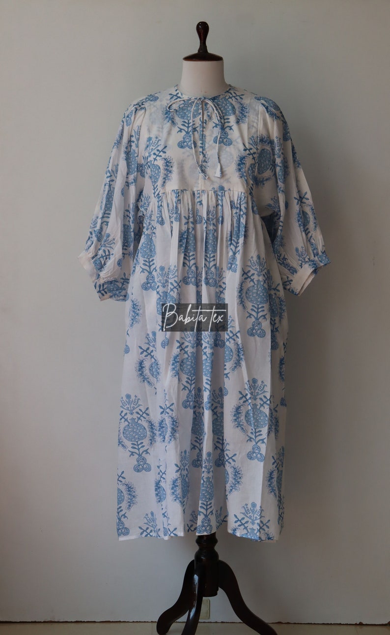 May include: A white cotton dress with a blue floral print and a tie at the neck. The dress has a gathered waist and 3/4 sleeves. The dress is on a mannequin.