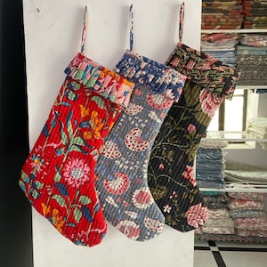 May include: Four quilted Christmas stockings with floral patterns hang against a white wall. The stockings feature red, gray, and black backgrounds with colorful flower designs. Each stocking has a ruffled top and a loop for hanging.